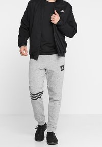 Mustakasta vetoketjullinen pommittaja-takki mustan t-paidan päällä, yhdistettynä vaaleanharmaisiin verkkohousuihin, joissa on musta logo ja kolme mustaa raitaa lahkeiden lähellä.