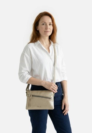 Vrouw met rood haar, gekleed in een witte blouse en donkere spijkerbroek, houdt een beige leren schoudertas met een textuur en een voorvak met ritssluiting vast.