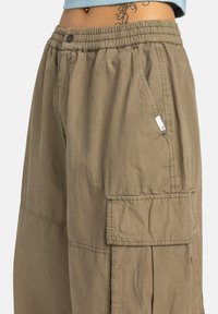 Zbliżenie luźno dopasowanych khaki spodni cargos z elastycznym paskiem i boczną kieszenią, noszonych przez osobę z tatuażem na talii.