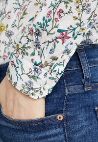 Main dans la poche d'un jean bleu avec un poignet de chemise blanche à manches longues présentant un imprimé floral multicolore et des boutons blancs.