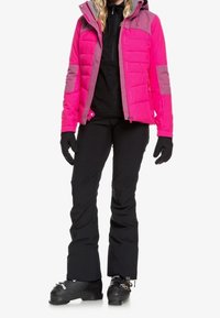 Giacca isolante rosa brillante con dettagli grigi, guanti neri, pantaloni neri e scarponi da sci. Presenta un cappuccio e una texture trapuntata.