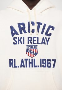 Sweat à capuche blanc avec texte bleu marine "ARCTIC SKI RELAY" et logo bouclier rouge, blanc et bleu avec l'inscription "POLO USA" au-dessus du texte "RL. ATHL. 1967".