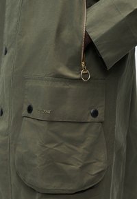 Κοντινό πλάνο στην πράσινη τσέπη του μπουφάν Barbour με μαύρα κουμπιά και ένα χρυσό φερμουάρ στη ραφή της πλευράς.