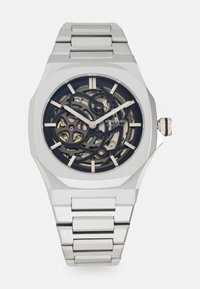 Montre en acier inoxydable argenté avec un cadran squeletté, mettant en valeur des engrenages complexes sur un fond noir. Dotée d'un boîtier géométrique et d'un bracelet à maillons.