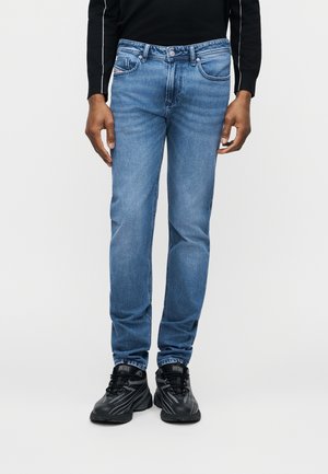 Mees, kes kannab helehalli sirge lõikega teksapükse, musti tossusid ja musta pika varrukaga pluusi, taustaks on lihtne taust.