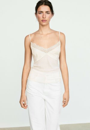 Ung kvinde iført en hvid blonderkantet camisole og hvide bukser står mod en ensfarvet lys baggrund.