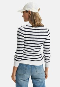 Pull en coton à côtes avec des rayures horizontales bleues marine et blanches, col rond et poignets ajustés, associé à un jean bleu et une casquette blanche.