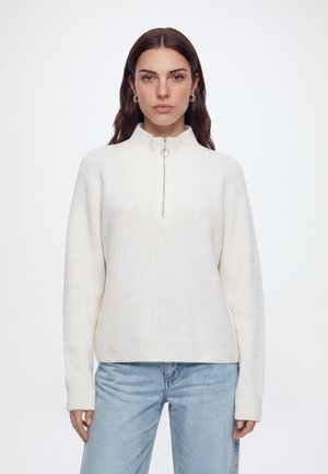 Femme aux longs cheveux foncés portant des boucles d’oreilles en or, un pull à demi-zip en maille côtelée blanche et un jean bleu clair, debout devant un fond blanc uni.