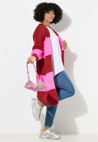 Cardigan ai ferri colorato con strisce rosa e rosse, camicia bianca, jeans blu e sneakers argento. Tiene una borsa rosa scintillante.
