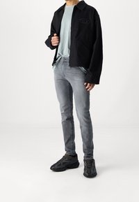 Svart jacka med logotyp, ljusblå t-shirt, grå slimfit-jeans och svarta texturerade sneakers, stående mot en vit bakgrund.