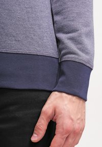 Felpa grigia con un corpo testurizzato e polsini a costine blu navy. Presenta cuciture a punto e una vestibilità aderente sui polsi.