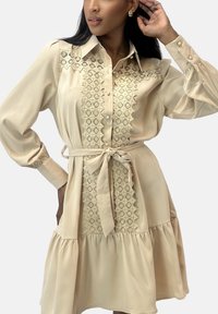 Beige jurk met een knoopsluiting aan de voorkant, kanten details langs het lijfje, lange mouwen en een strik in de taille, met een uitlopende rok.