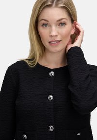 Blondynka z niebieskimi oczami, ubrana w fakturowaną czarną kurtkę z srebrnymi guzikami, dotyka prawego ucha na białym tle.