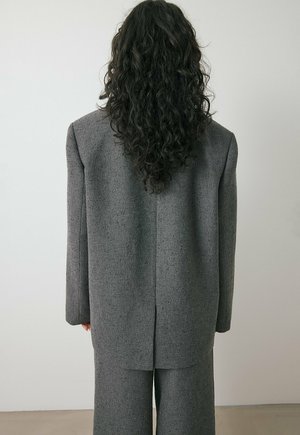 Person with long curly dark hair wearing a textured gray blazer and matching wide-leg pants, standing against a plain light wall.