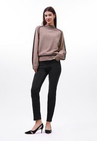 Beige kötött pulóver fekete részletekkel, hosszú ujjaival és bordázott mandzsettával; fekete skinny farmernadrággal és fekete magassarkú cipővel párosítva.
