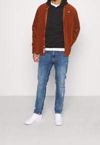 Camisa de botón de pana marrón sobre una sudadera negra con capucha y una camiseta blanca, combinada con jeans desgastados azules y zapatillas blancas.
