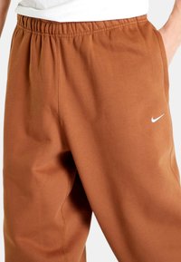 Nike-housut ruskeasta kankaasta, jossa on elastinen vyötärö, sivutaskut ja pieni valkoinen logo vasemmassa lahkeessa. Sujuva pinta.