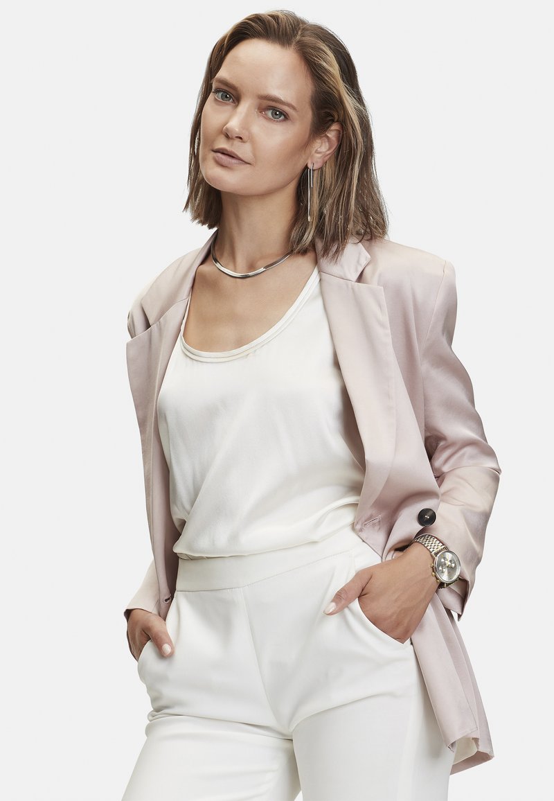 Blazer en satin rose pâle sur une blouse blanche, associé à un pantalon blanc ajusté. Accessoirisé avec un collier et une montre en argent.