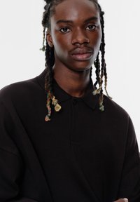 Zwarte poloshirt met een kraag en een enkele knop. Heeft een losse pasvorm en een gladde textuur. Opvallende kleurrijke gevlochten haaraccenten.