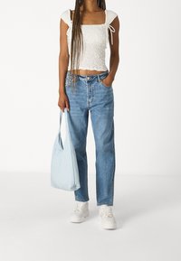 Mujer vestida con un top corto blanco con fruncidos y jeans de tiro alto azul, sosteniendo una bolsa de mano azul clara y usando zapatillas blancas, sobre un fondo blanco.