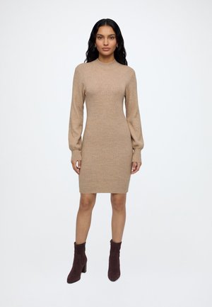 Femme debout portant une robe en maille côtelée beige à manches longues et des bottines marron foncé sur un fond blanc uni.