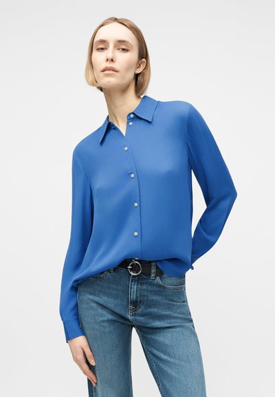 Γυναίκα με μπλε μπλούζα μακριών μανικιών με κουμπιά, φορεμένη μέσα σε μπλε τζιν και μαύρη ζώνη, πάνω σε απλό λευκό φόντο.