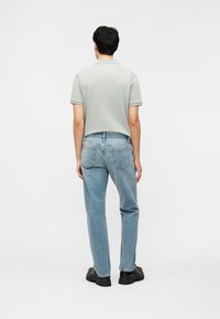 Άνδρας που στέκεται γυρισμένος με την πλάτη, φοράει ανοιχτό γκρι πόλο μπλουζάκι, ανοιχτό μπλε τζιν και μαύρα ογκώδη παπούτσια σε λευκό φόντο.