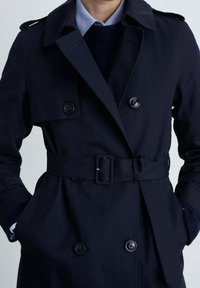 Osoba oblečená v námořnickém modrém dvouvrstvém trenčkotě s páskem v pase, ramenními nášivkami a rukama v kapsách, pod modrou košilí a svetrem.