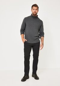 Dunkelgrauer gestrickter Rollkragenpullover, schwarze Slim-Fit-Hose und schwarze Schnürboots. Das Modell steht selbstbewusst mit den Händen in den Taschen.
