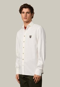 Camisa blanca de manga larga y cuello con botones marrones y un logo bordado en el lado izquierdo del pecho.
