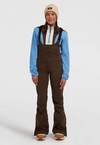 Frau in braunen Schneebib-Pants, blauer und brauner Zip-Jacke, beiger Strickmütze und beigen Winterstiefeln, die vor einem weißen Hintergrund steht.