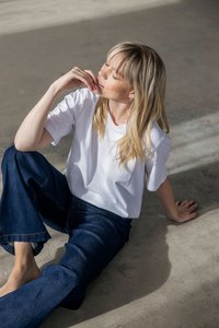 Mlada žena s dugom plavom kosom sjedi bosa na betonskom podu, nosi bijelu majicu i plave traperice, dodiruje usta rukom.