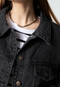 Giacca di jeans nera con colletto a punta e bottoni frontali, indossata sopra una camicia bianca con righe nere; collana d'oro semplice visibile.