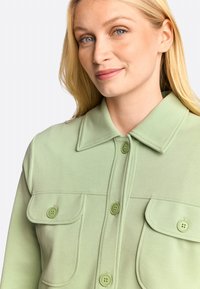 Light green button-up shirt with a collar, featuring two chest pockets and matching buttons. Made from a smooth fabric with a slight sheen.