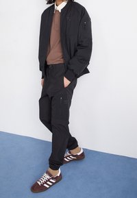 Černá bomber bunda, hnědý polo, černé cargo kalhoty. Bordó a bílé tenisky mají tři pruhy. Hladký materiál, pohodlný střih, funkční kapsy.