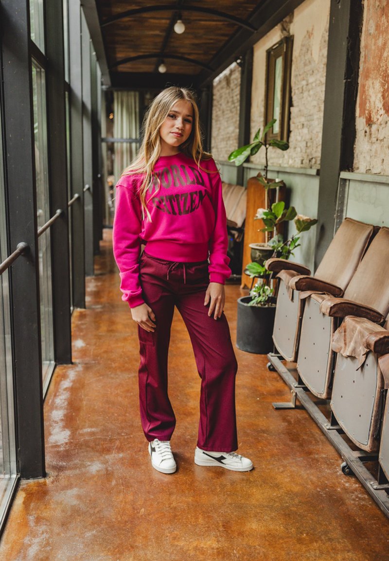 Roze sweatshirt met zwarte tekst "WORLD CITIZEN", gecombineerd met bordeaux brede broek. Witte sneakers met zwarte accenten. Binnenomgeving.