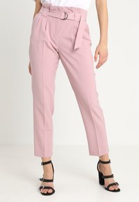 Pantalons rose clair à taille haute, ceinture avec un anneau en argent, plis à l'avant et design à jambes fuselées. Portés avec des talons noirs à lanières.