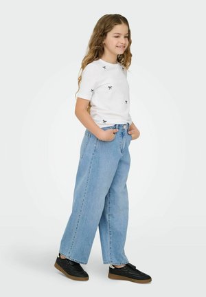 Jente med langt, bølgete hår som har på seg en hvit t-skjorte med små sløyfetrykk, lyseblå vide jeans og svarte joggesko, hendene i lommene.