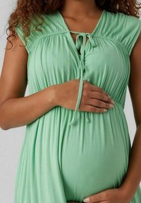 Mujer embarazada vestida con un vestido sin mangas de color verde claro, sosteniendo suavemente su barriga con ambas manos.