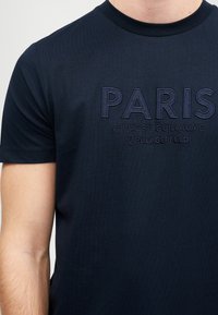 Navy zila T-kreklu ar izšūtu tekstu "PARIS 21 RUE ST-GUILLAUME KARL LAGERFELD" uz krūts.