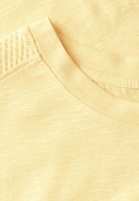 Krátký rukáv světle žluté tričko s texturovaným výstřihem, hladkým materiálem a jemnými variacemi v textuře po celém povrchu.