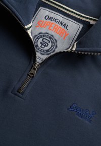 Granatowa bluza z suwakiem wykonana z miękkiego materiału, posiada wysoki kołnierz, haftowane logo oraz kontrastową metkę z nazwą marki i emblematem.