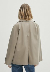 Olkapäihin ulottuvat vaaleat hiukset omaava nainen, jolla on päällään löysä beige takki ja siniset farkut, kuvattuna selin yksinkertaisen taustan edessä.