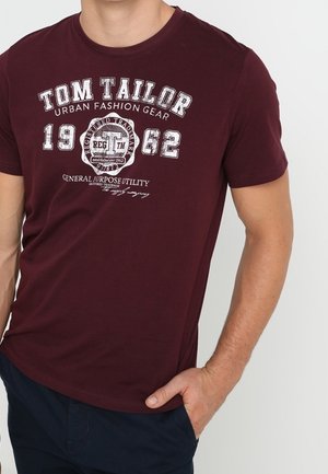 Bordeauxfarbenes Tom Tailor T-Shirt mit weißem Text und Logo, getragen von einer Person, die eine Hand in der Tasche einer marineblauen Hose hat.