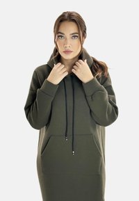 Vestido con capucha en verde olivo de tejido suave. Cuenta con un gran bolsillo delantero, capucha con cordón y un corte holgado con mangas largas.