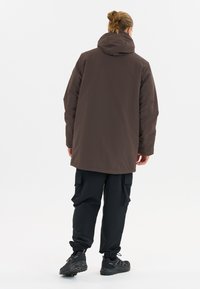 Braune wasserdichte Jacke mit Kapuze, im lässigen Schnitt und mit überschnittenen Schultern. Kombiniert mit schwarzen Cargo-Hosen und robusten schwarzen Schuhen.