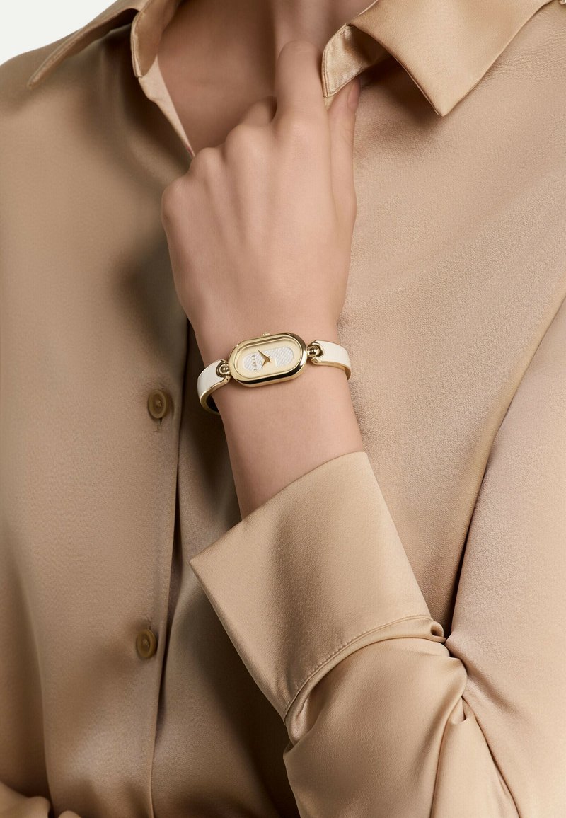 White strap watch with a gold oval case and textured cream dial. Worn on a hand, paired with a beige satin shirt.