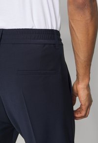 Marineblauwe broek met een elastische tailleband, voorzien van een zijzak en een platte achterzak. Gladde stofstructuur, rechte broekspijpen.