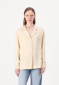 Lys beige skjorte med knappestolpe og en teksturert, subtil bladmønster. Har klassisk krage og lange ermer med knappefeste.