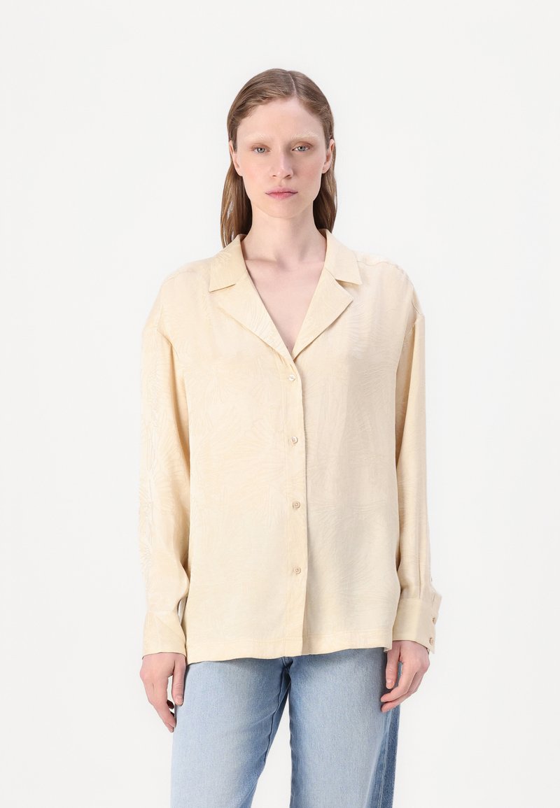 Lys beige skjorte med knappestolpe og en teksturert, subtil bladmønster. Har klassisk krage og lange ermer med knappefeste.